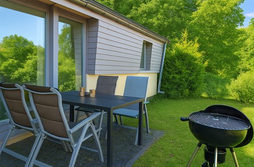 Unser Ferienhaus im Ferienpark Eifel Terrasse vor dem Ferienhaus mit Tisch und Stühlen, Grill und Blick auf grünes Umland.