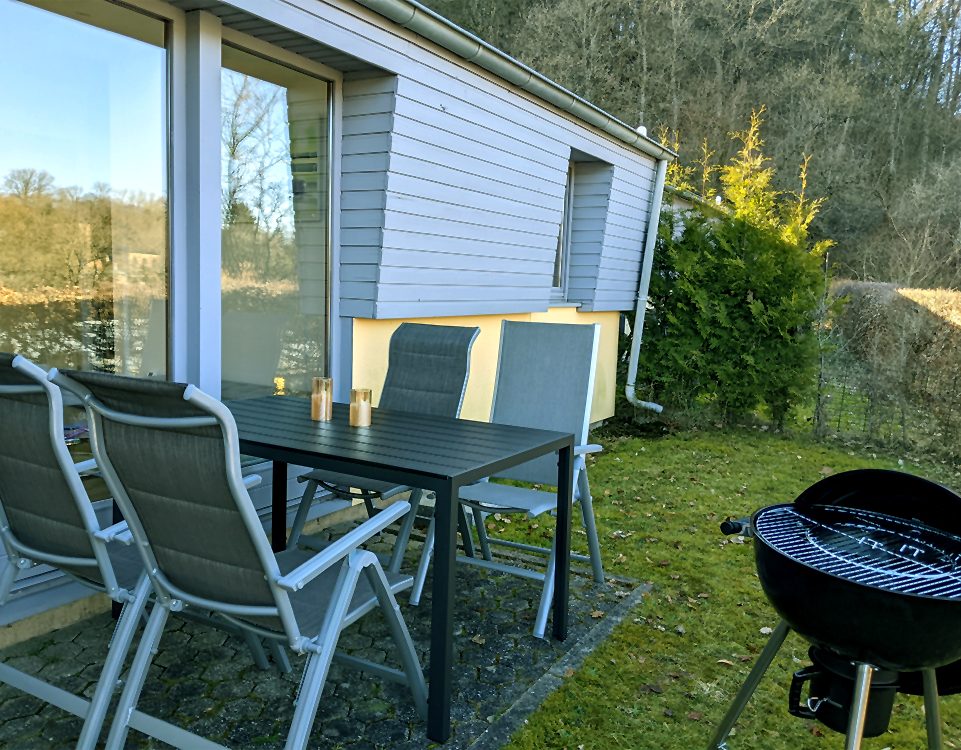Ferienhaus - Terrasse Terrasse mit Tisch, Stühlen und Grill vor dem Haus, umgeben von grüner Landschaft.